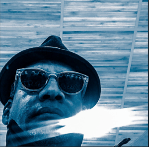 Image of Saint Woke, also known as Vince Warren, the Executive Director of the Center for Constitutional Rights. He is wearing a hat and sunglasses, looking down at the camera. The image is tinted blue.