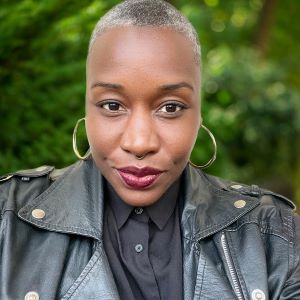 A phot of Sunyata Altenor, the new Communications Director. She is wearing a leather jacket with a black buttom-up shirt underneath. She has gold hoop earrings on and dark red lipstick. She is looking directly at the camera. Behind her are green plants.
