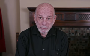 Picture of Bill Goodman, he is sitting in a chair, visible from the chest up. He is staring directly at the camera and is wearing a black button-up shirt. A fireplace mantle is visible behind him.