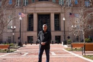 Plaintiff Salah Al-Ejaili, outside the courthouse in Alexandria, Virginia in March. He is wearing glasses, a black jacket, and he is standing with his hands crossed in front of him. There are two U.S. flags flying on either side of the courthouse behind him.