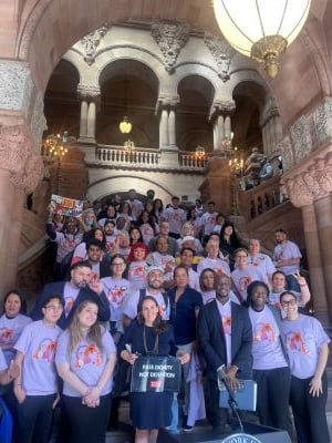 Staff Attorney Samah Sisay is pictured here, second from the right in the front row, with our partners in Albany. There is a large group of people up the steps of the capitol building in Albany. They are wearing matching t-shirts. Many people are smiling.
