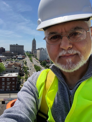 selfie of our cleint kifah jayyousi wearing a construction hat and vest. he is wearing glasses and looking at the camera
