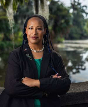 Photo of Colette Pichon Battle standing in front of a bayou. She is wearing a black blazer with a green shirt underneath, and a shell necklace. She has her arms crossed in front of her and is looking directly at the camera.