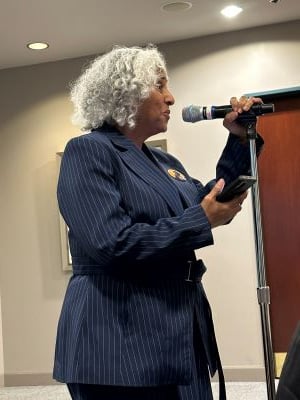 A side photo of our board member and Jackson water partner Makani Themba standing and speaking at a microphone. She is wearing a blue pin stripe jacket. She is holding the mic in one hand and her phone in her other hand.