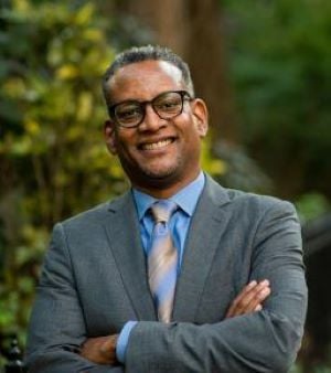 Photo of our Executive Director Vince Warren. He has his arms crossed in front of him and he is smiling directly at the camera. He is wearing a grey suit, with a blue shirt underneath and a blue and gold tie. He has thick black rimmed glasses. Green trees are behind him.