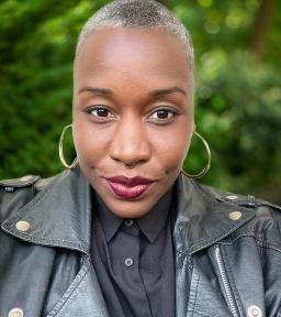 Headshot of Sunyata with silver hoop earrings and a black motorcycle jacket in front of a mottled green background