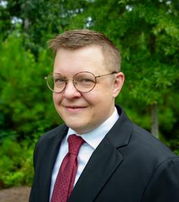 Headshot of CJ Sandley in navy blazer, white shirt, and red tie in front of green trees