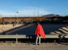 Lone man wrapped in a red blanket stands before a border wall
