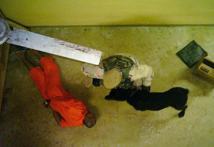A prisoner with his hands handcuffed behind his back lies on the floor while a guard with a dog stands over him 