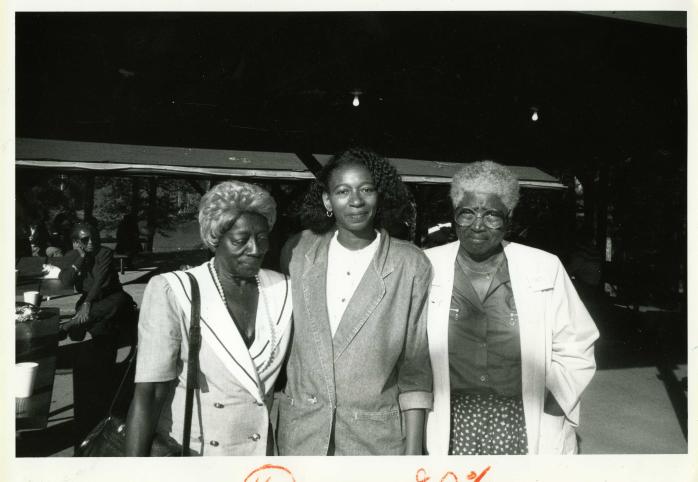 Three Black women posing for a photo. One is smiling while looking down and the other two are smiling at the camera.