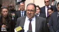 This is a screengrab from a press conference. Baher Azmy is speaking to press, he is wearing a dark gray suit, white button down, brown tie and has glasses on. His hair is dark and cut short.