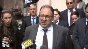 This is a screengrab from a press conference. Baher Azmy is speaking to press, he is wearing a dark gray suit, white button down, brown tie and has glasses on. His hair is dark and cut short.