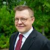 Headshot of CJ Sandley in navy blazer, white shirt, and red tie in front of green trees